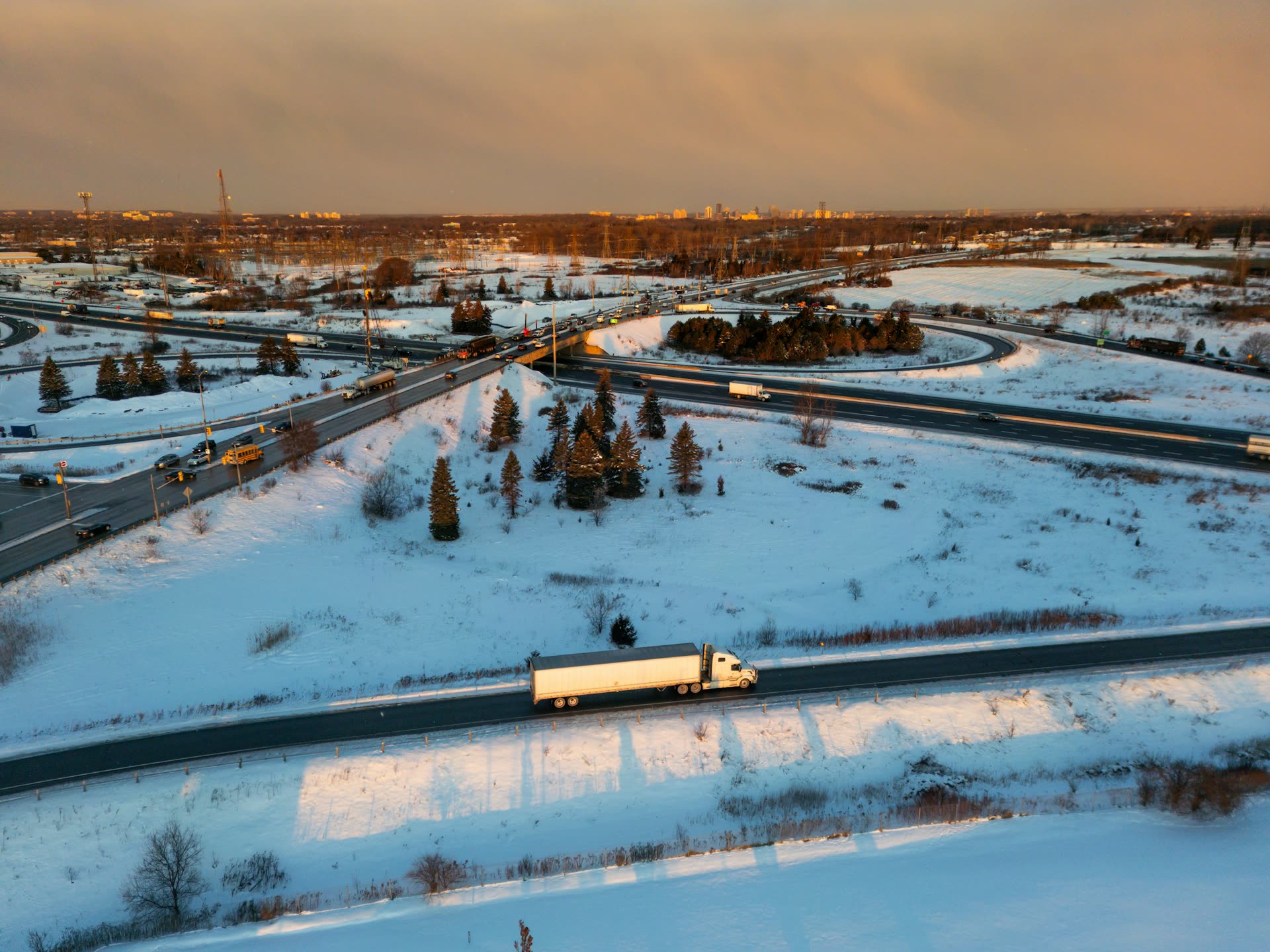 A tractor-trailer on a snow-lined highway at dusk, with an interchange and a city skyline behind it